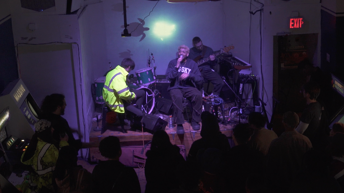 Band performing in a dark room with people around a stage, illuminated by a bright light.